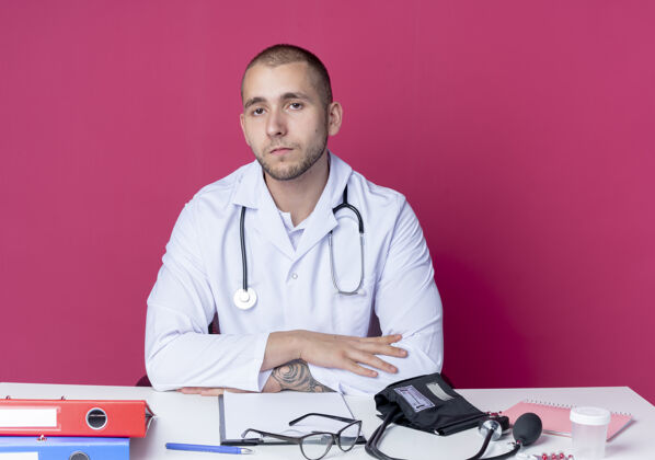 人面容严肃的年轻男医生穿着医用长袍和听诊器坐在办公桌旁 手放在办公桌上 看着隔离在粉红色墙上的前面医生脸坐着