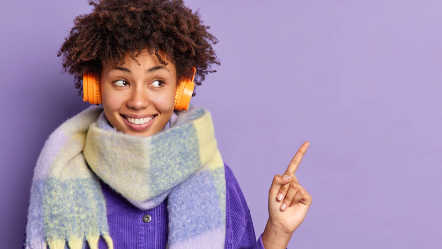 手指年轻的非洲裔美国妇女戴着耳机 脖子上戴着暖和的围巾 感觉很高兴 指着复印空间耳机乐趣指向