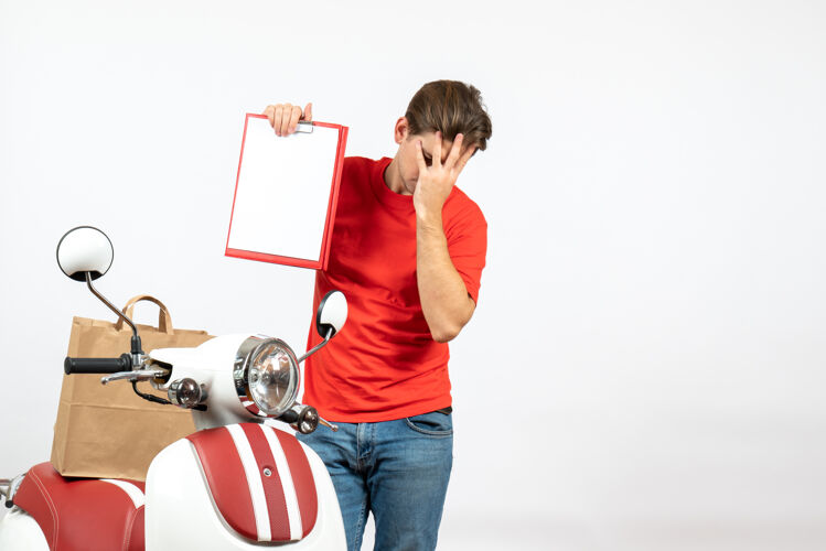 情绪顶视图年轻情绪过度疲惫的送货员身穿红色制服 站在滑板车旁 在白色墙上展示文件微笑年轻人人