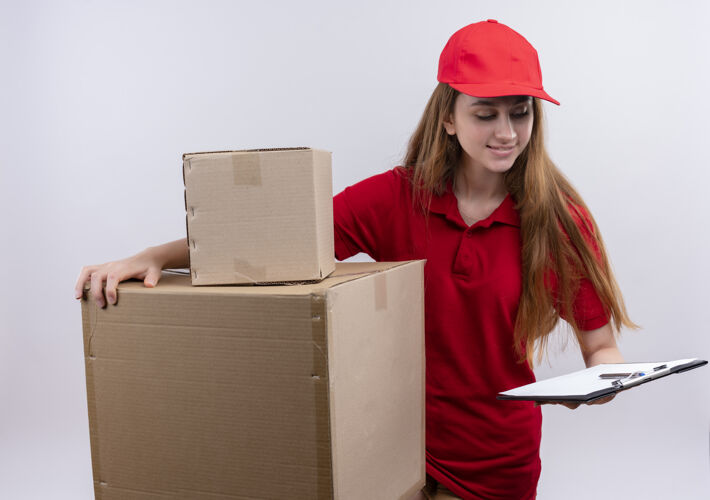 微笑微笑着的年轻送货女孩穿着红色制服拿着盒子和剪贴板 看着剪贴板上孤立的空白递送女孩盒子
