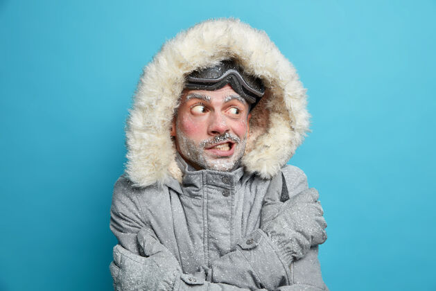 成人一张欧洲男子在溜冰后因寒冷而发抖的照片 他双手交叉着身体试图取暖 穿着灰色的冬季夹克 带着毛皮兜帽和手套 冻得满脸都是冰惊艳站立人