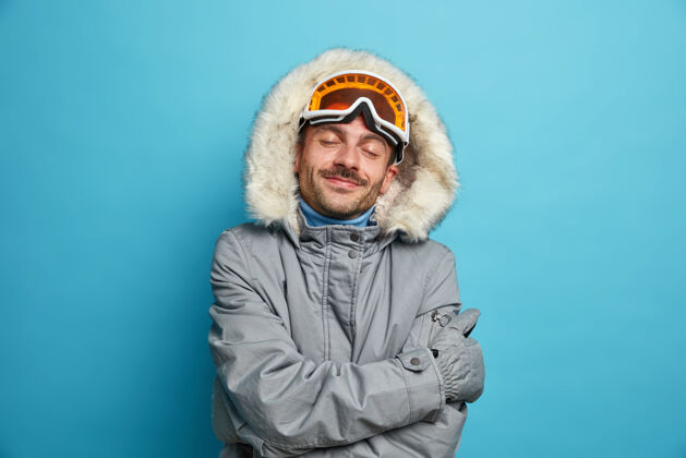 蓝色高兴的男子滑雪板感觉舒适和温暖的冬季夹克拥抱自己回忆美好的时刻去滑雪在寒冷的天气里站着闭上眼睛爱好运动装冬季假期