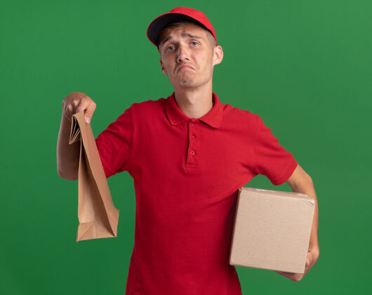 男孩失望的年轻金发送货男孩拿着纸包装和卡片盒在绿色失望金发包装