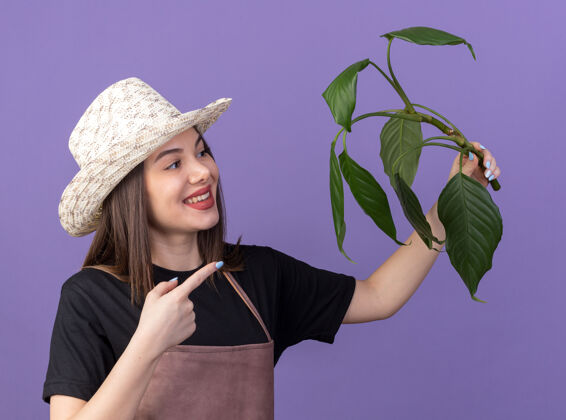 树枝美丽的高加索女园丁微笑着戴着园艺帽 看着并指着紫色的植物枝桠帽子漂亮指尖