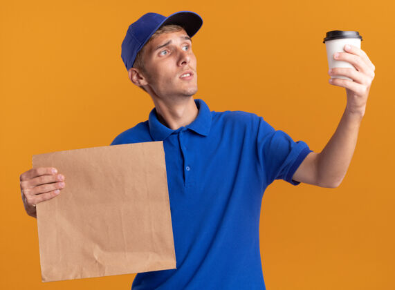 送货困惑的年轻金发送货男孩拿着纸包裹 看着橙色的杯子金发杯子年轻