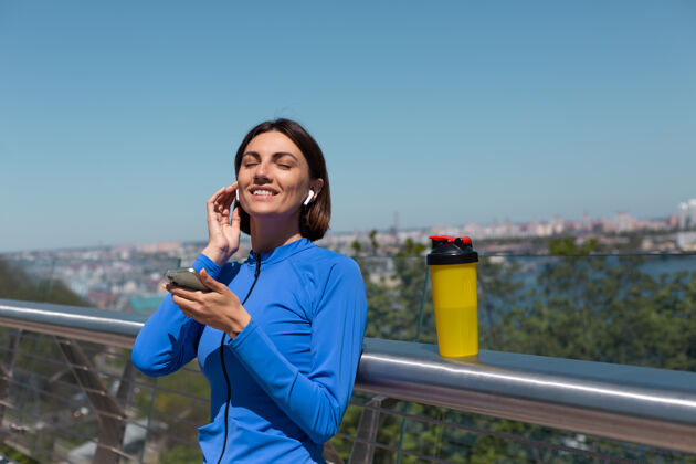 运动员穿着蓝色运动服的年轻女子 在阳光明媚的早晨 戴着无线耳机和手机 在桥上休息 听音乐锻炼女性自信