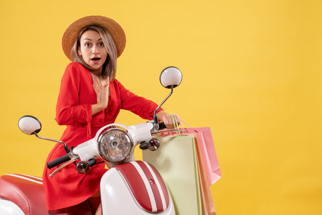 车一个穿着红色衣服的金发女人坐在轻便摩托车上 拿着购物袋指着自己金发女人抱着人