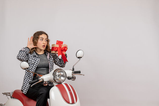 微笑前视图好奇的年轻女子骑着轻便摩托车 手里拿着礼物 听着灰色墙壁上的东西肖像轻便摩托车人