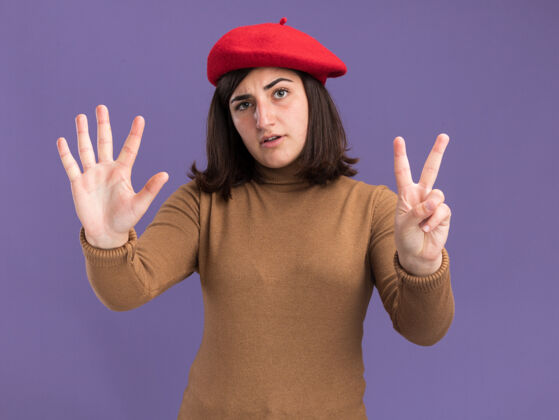 手势自信的年轻漂亮的白人女孩戴着贝雷帽 七个手指在紫色上做手势贝雷帽帽子七