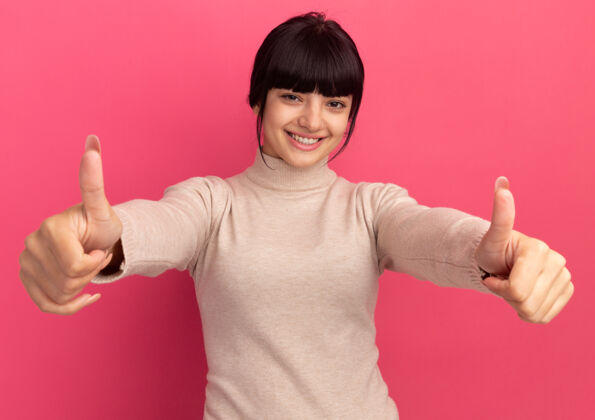 手年轻的黑发白人女孩微笑着竖起大拇指 双手放在粉红色的衣服上黑发拇指年轻