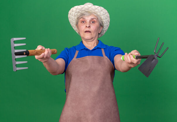女印象深刻的上了年纪的女园丁戴着园艺帽拿着耙子和锄头耙在草地上老人印象园艺