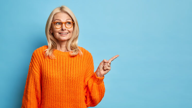 中间体贴的金发四十岁的欧洲女人戴着眼镜和针织橙色毛衣指着复印空间人眼镜离开