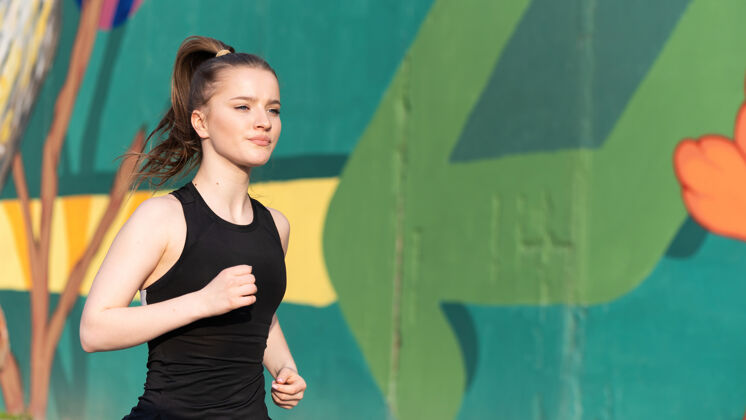 跟踪身着运动服的金发女郎在户外训练的路上奔跑 背景是五彩缤纷的墙壁手健身护理