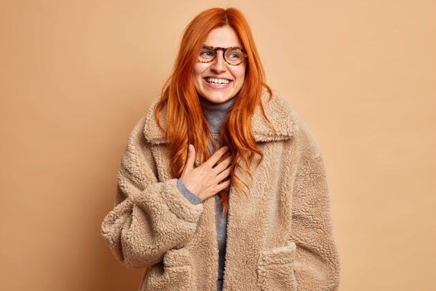 微笑喜出望外的红发成年女子笑着表达快乐真挚的情感戴着眼镜和温暖的棕色皮草外套聚精会神地笑着享受冬天的时光有着乐观的心情时尚的概念米色大笑正面