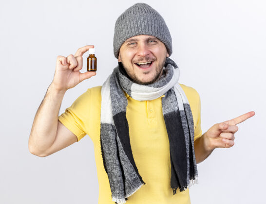 瓶子快乐的年轻金发病斯拉夫男子戴着冬季帽子和围巾拿着在玻璃瓶中的药指着隔离在白色墙壁与复制空间的一面指尖举行疾病