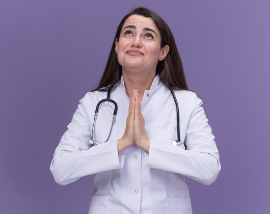焦虑焦急的年轻女医生穿着带听诊器的医用长袍 双手合十 仰望紫色医学女一起