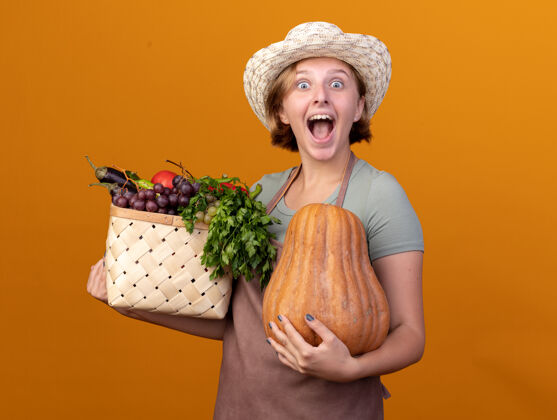 蔬菜兴奋的年轻斯拉夫女园丁戴着园艺帽 手里拿着菜篮子和橘子上的南瓜南瓜年轻花园