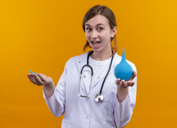 听诊器快乐的年轻女医生穿着医用长袍 手持听诊器在孤立的橙色空间灌肠长袍医疗女性