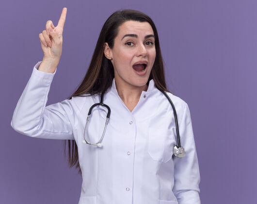 惊喜惊讶的年轻女医生穿着医用长袍用听诊器指着紫色长袍点医生