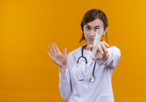 手势严肃的年轻女医生穿着医用长袍和听诊器伸展注射器和手势停止在孤立的橙色空间与复制空间停止医生穿着
