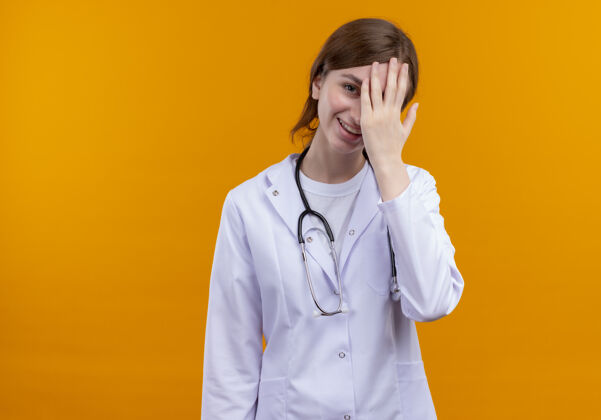 年轻笑着的年轻女医生穿着医用长袍和听诊器把手放在脸上孤立的橙色空间与复制空间医学医生脸