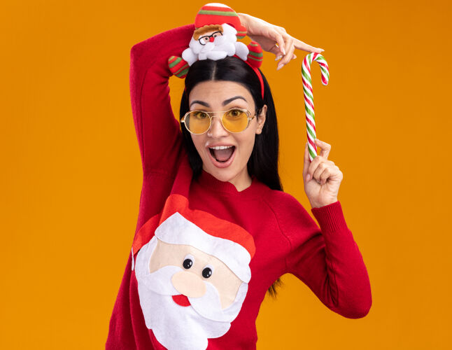眼镜令人印象深刻的年轻白人女孩戴着圣诞老人的头带和毛衣 戴着眼镜 手持传统的圣诞糖果手杖 靠近头部 看着隔离在橙色背景上的相机女孩毛衣橘子
