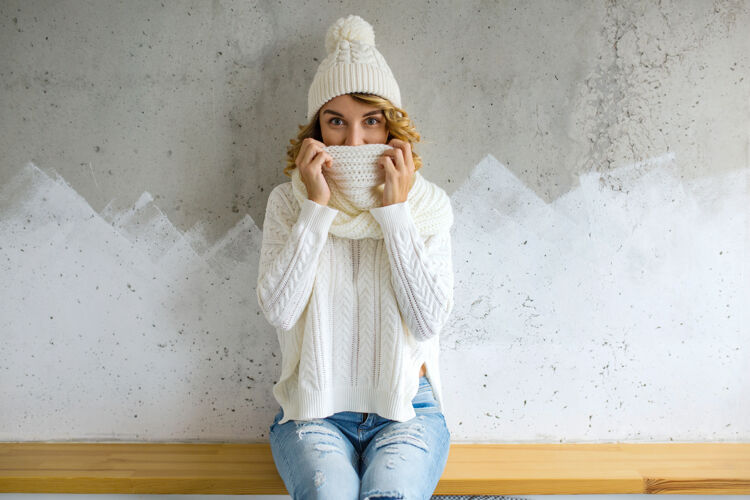 室内漂亮的年轻女子靠墙坐着 穿着白毛衣 戴着针织帽子和围巾休息坐着圣诞节