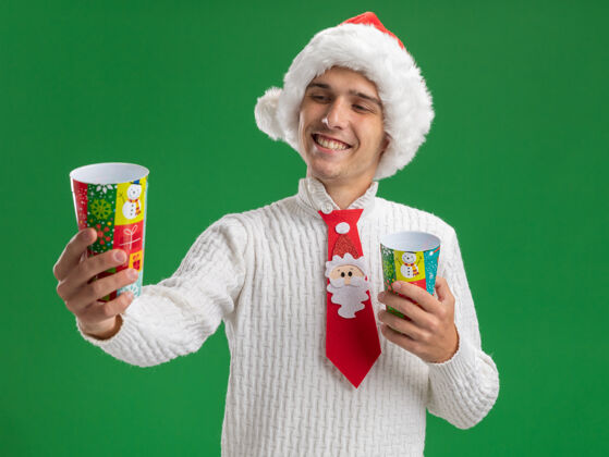 举行快乐的年轻帅哥戴着圣诞帽 打着圣诞老人的领带 拿着塑料圣诞杯 伸着懒腰 看着其中一个孤立的绿色背景圣诞老人绿色领带
