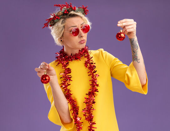 周围困惑的金发女郎头戴圣诞花环 脖子上戴着金箔花环 戴着眼镜 手里拿着圣诞饰品 看着其中一个孤立的紫色背景脖子紫色眼镜