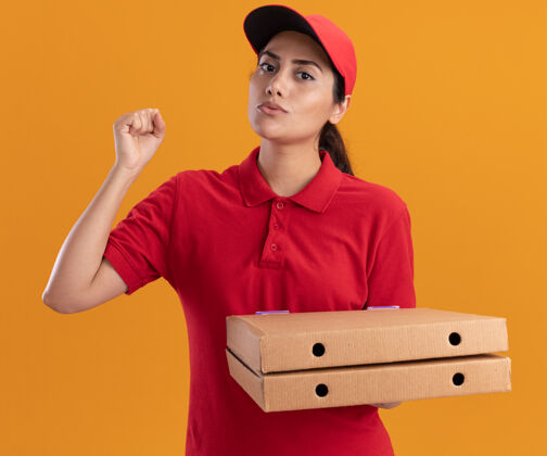 制服严格的年轻送货女孩穿着制服 戴着帽子拿着披萨盒举起拳头隔离在橙色的墙上脸帽子人