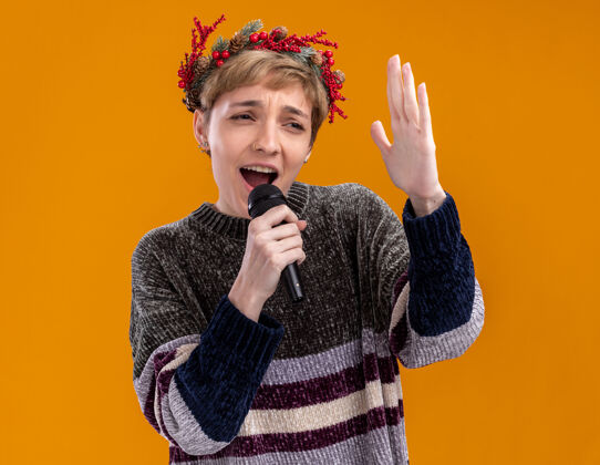 花环年轻漂亮的女孩戴着圣诞花环 拿着麦克风近嘴 手举在空中 看着一边唱歌 在橙色背景下孤立圣诞快乐橙色嘴