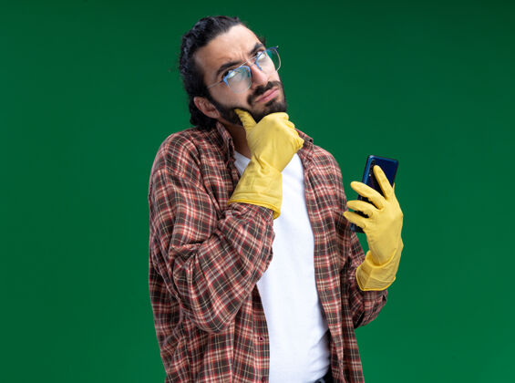 帅哥迷茫的年轻帅气的清洁工穿着t恤 戴着手套拿着电话 把下巴孤立在绿色的墙上电话手套抓