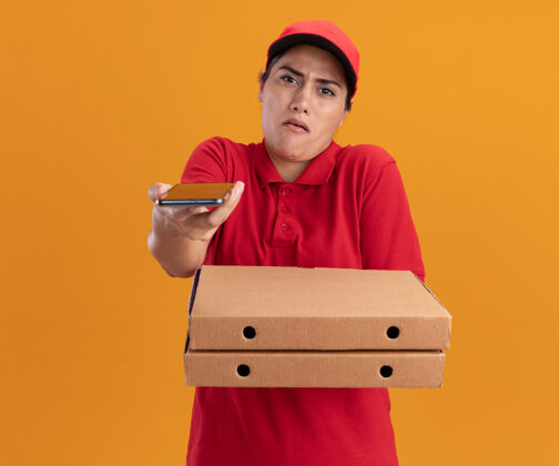 感觉一个穿着制服 戴着帽子 拿着披萨盒 拿着电话的年轻送货员站在橙色的墙上表情人电话