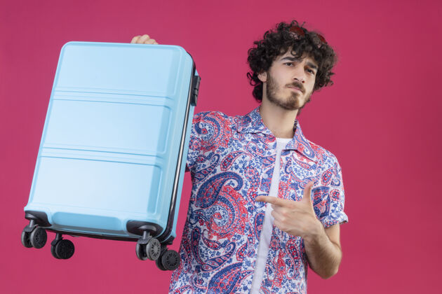 自信自信的年轻帅气的卷发旅行者头戴太阳镜拿着手提箱指着孤立的粉色空间穿手提箱抱着