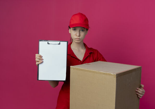 纸箱自信的年轻漂亮的送货女孩 穿着红色制服 戴着帽子 拿着纸盒 在深红色的背景上展示独立的剪贴板 还有复印空间盒子漂亮制服
