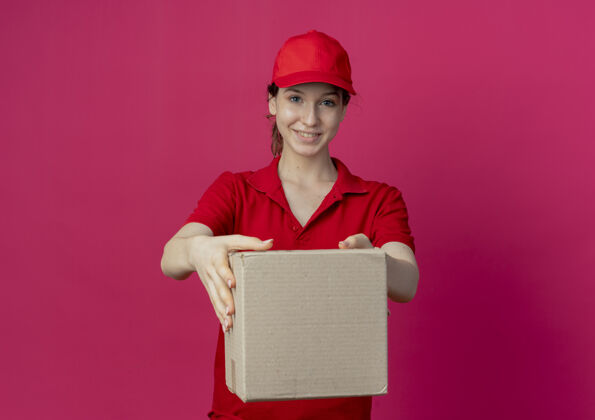 制服微笑着的年轻漂亮的送货女孩 穿着红色制服 戴着帽子 在摄像机前伸出纸箱 在深红色的背景下与复印空间隔离开来伸展红色背景