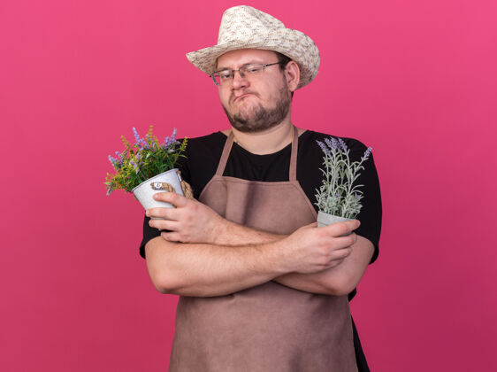 信心自信的年轻男园丁戴着园艺帽 在粉红色墙上的花盆里拿着和交叉的花帽子穿着姿势