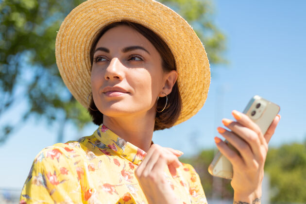 肖像在阳光明媚的日子里 穿着黄色夏装戴着帽子的女人的户外贴身写真拿着手机往左看科技模型休闲