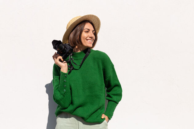 博客穿着绿色休闲毛衣和帽子的女士户外白墙上快乐的正面游客带着专业相机女性墙成人
