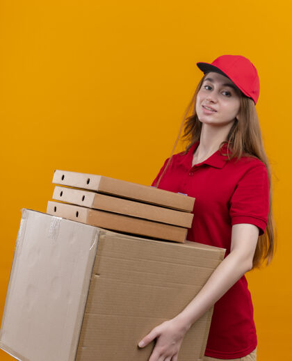 制服自信的年轻送货女孩穿着红色制服 把箱子和包裹放在孤立的橙色空间里信心女孩年轻