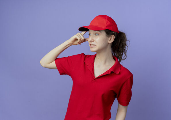 年轻体贴的年轻漂亮的送货女孩 穿着红色制服 戴着帽子 手指放在头上 看着紫色背景上孤立的一面 还有复印空间头壁板紫色