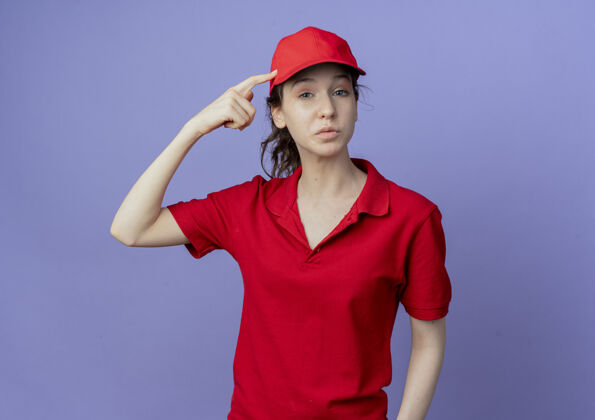 帽子体贴的年轻漂亮的送货女孩 穿着红色制服 戴着帽子 手指放在头上 紫色背景上 与世隔绝思想头年轻