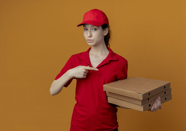漂亮令人印象深刻的年轻漂亮的送货女孩在红色制服和帽子举行 并指着比萨饼包装孤立的橙色背景与复制空间披萨帽子印象