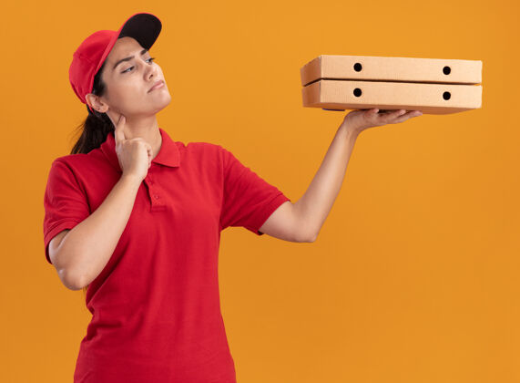 拿着想着穿着制服戴着帽子的年轻送货女孩 看着隔离在橙色墙上的比萨饼盒盒子年轻人感觉