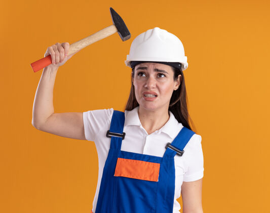 锤子不高兴地看着旁边穿着制服的年轻建筑妇女拿着锤子孤立地站在橙色的墙上脸女人人