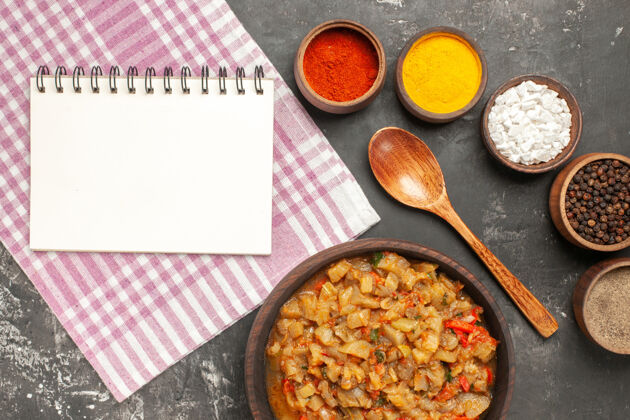 勺子顶视图烤茄子沙拉在碗木勺不同的香料在碗一个笔记本在黑暗的表面茄子沙拉一餐碗