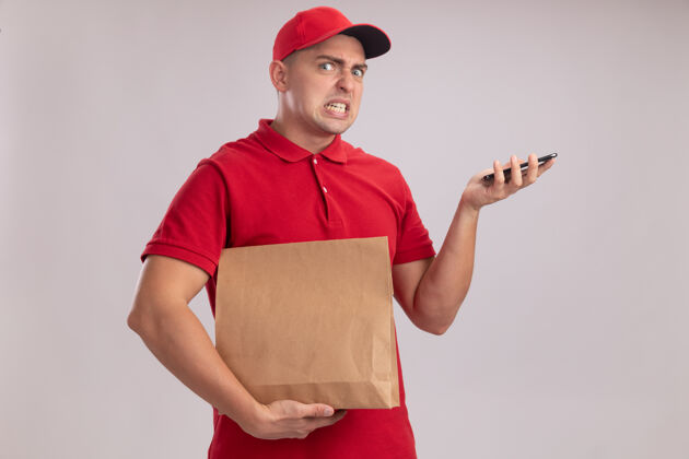 衣服愤怒的年轻送货员穿着制服 戴着帽子 拿着纸食品包 电话被隔离在白墙上制服姿势人