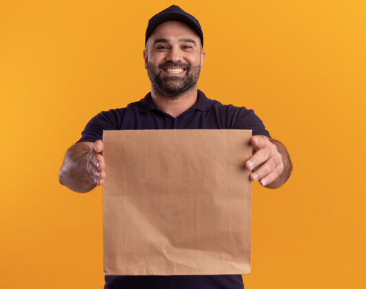 衣服微笑的中年送货员 身着制服 戴着帽子 在黄色的墙上隔着一个纸制食品包姿势食物制服