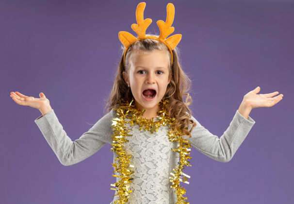 圣诞节兴奋的小女孩戴着圣诞发箍 脖子上戴着花环 双手分开放在蓝色背景上蓝色手花环