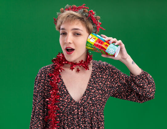 圣诞快乐好奇的年轻漂亮女孩戴着圣诞花环 脖子上戴着金属丝花环 耳边拿着塑料圣诞杯 听着秘密 看着隔离在绿色背景上的相机塑料花环秘密